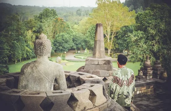 Homme se tenant à coté d'une statue de Bouddha dans un temple en pierre entouré de verdure