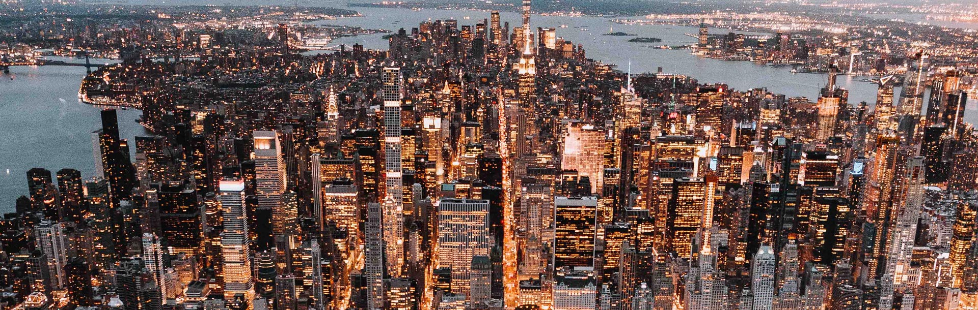 Vue aérienne de Manhattan illuminé à la tombée de la nuit, avec ses gratte-ciel scintillants, dont l’Empire State Building, s’élevant au cœur de l’île, bordée par les rivières Hudson et East River.