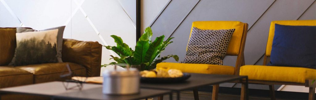 intérieur cosy d'une salle d'attente avec un canapé marron, des fauteuils jaune moutarde avec des coussins, une table basse et une plante verte