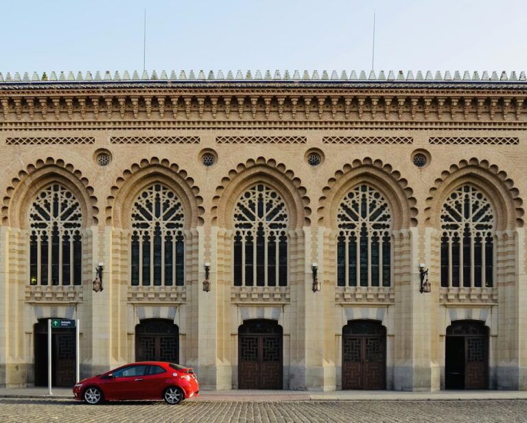 Voiture rouge garée devant un batiments sculptés, avec arcades et motifs