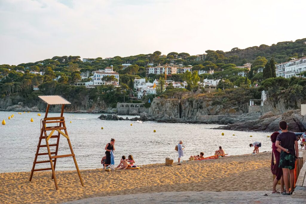 Plage de sabme blanc avec personnes, chaise de surveillance de baignade et côte avec habitations en hauteur et végétation