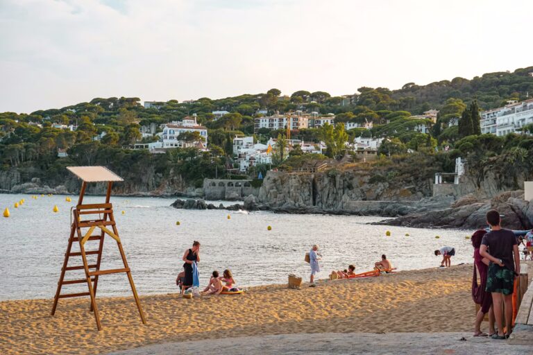 Plage de sabme blanc avec personnes, chaise de surveillance de baignade et côte avec habitations en hauteur et végétation