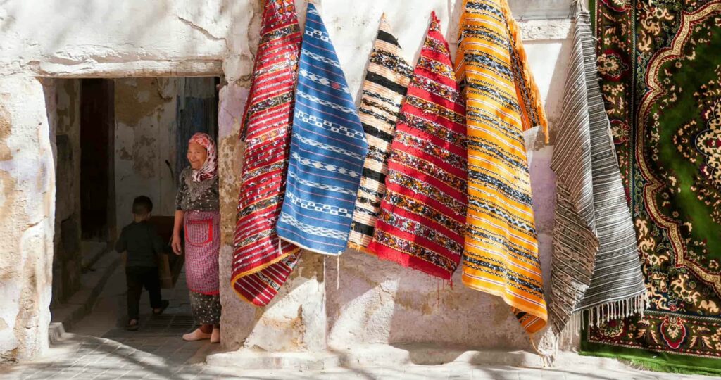 Mur de chaux recouvert de tapis colorés à vendre, une veille dame souriante à la porte, dans une rue de Fez, Maroc