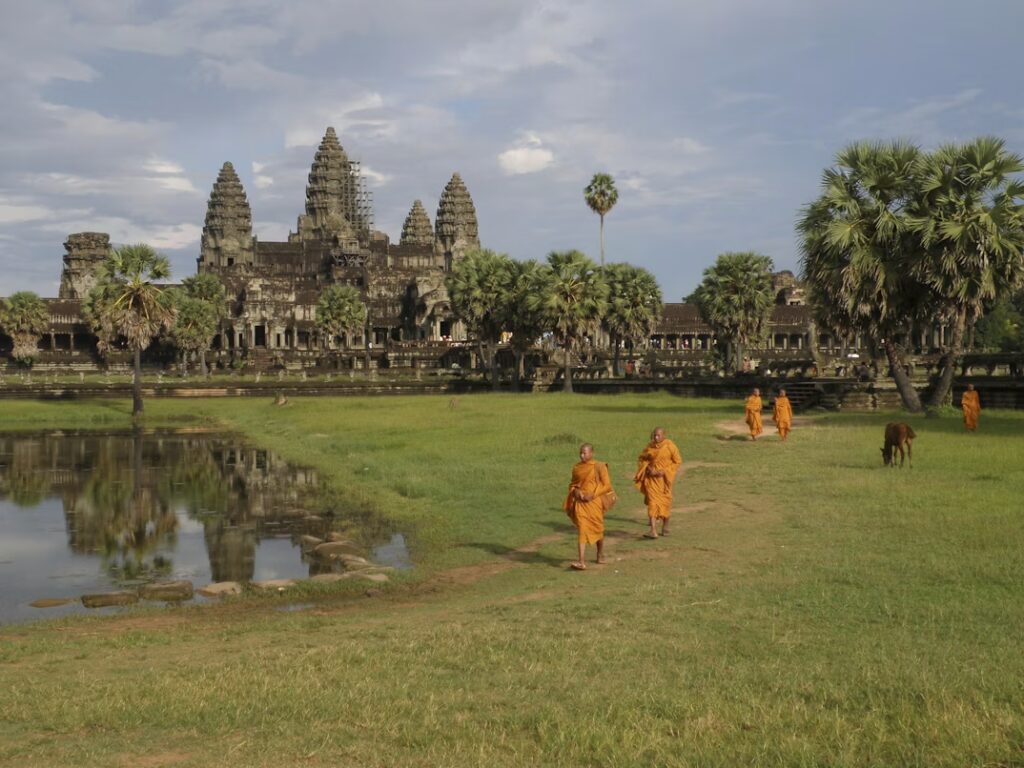 2tatendu d'herbe avec point d'eau et temples en pierre au fond, palmiers et moins en tenue traditionnelle jaune ocre