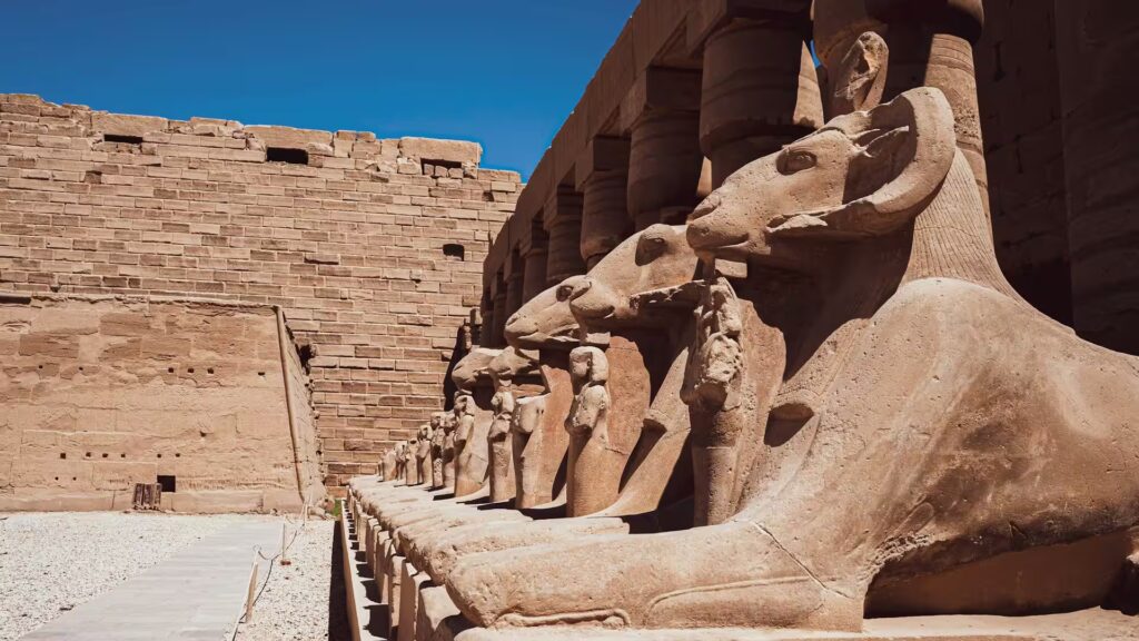 Rangée de statues à tête de belier avec des colonnes derriere et à droite d'un batiment en pierre