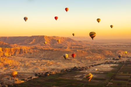 Vua arienne sur des montgolfieres dans le ciel au levé du soleil, au dessus de champs, ville et au loin des montagnes
